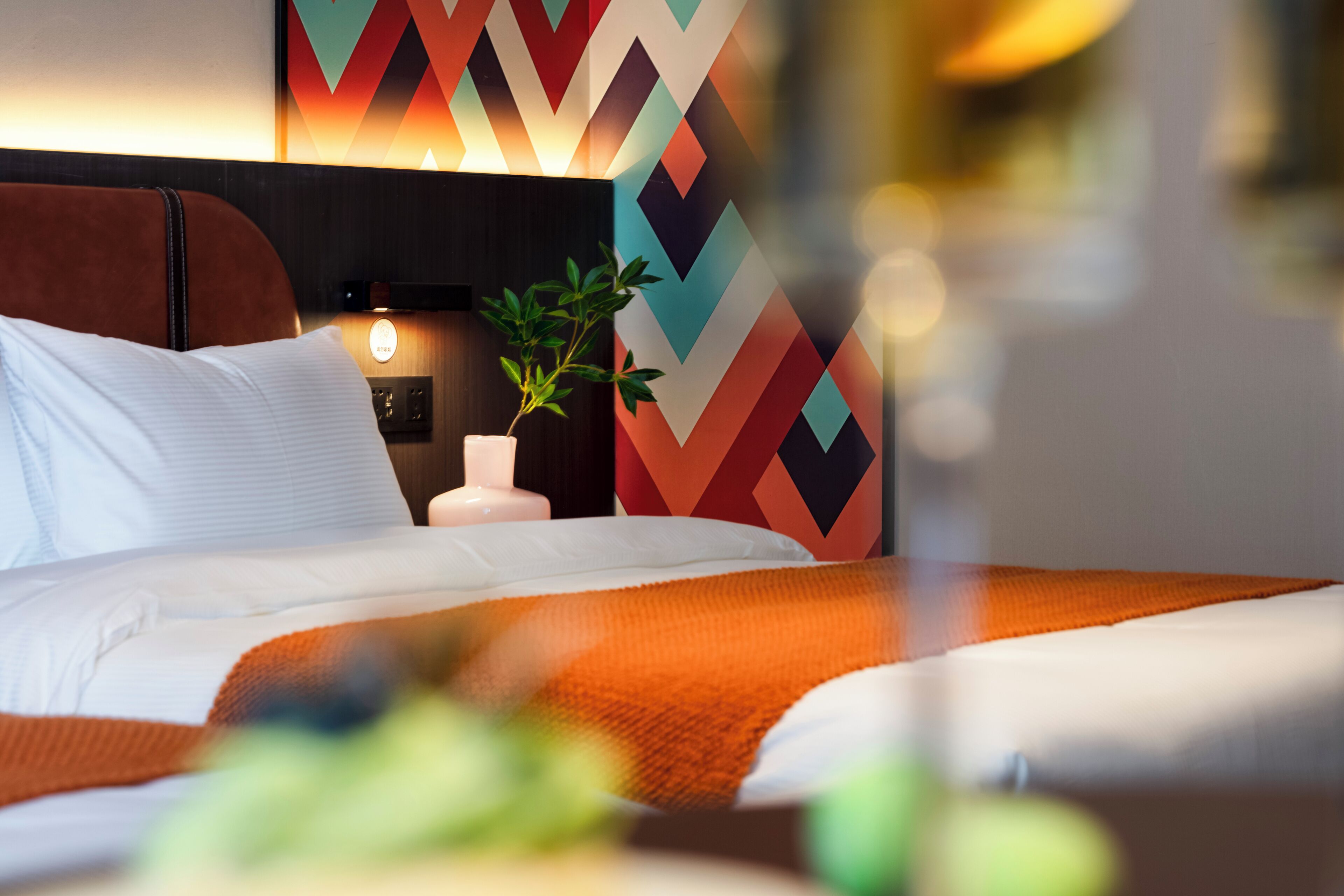 A modern hotel room with a bed, a nightstand, a lamp, and a decorative wall panel.