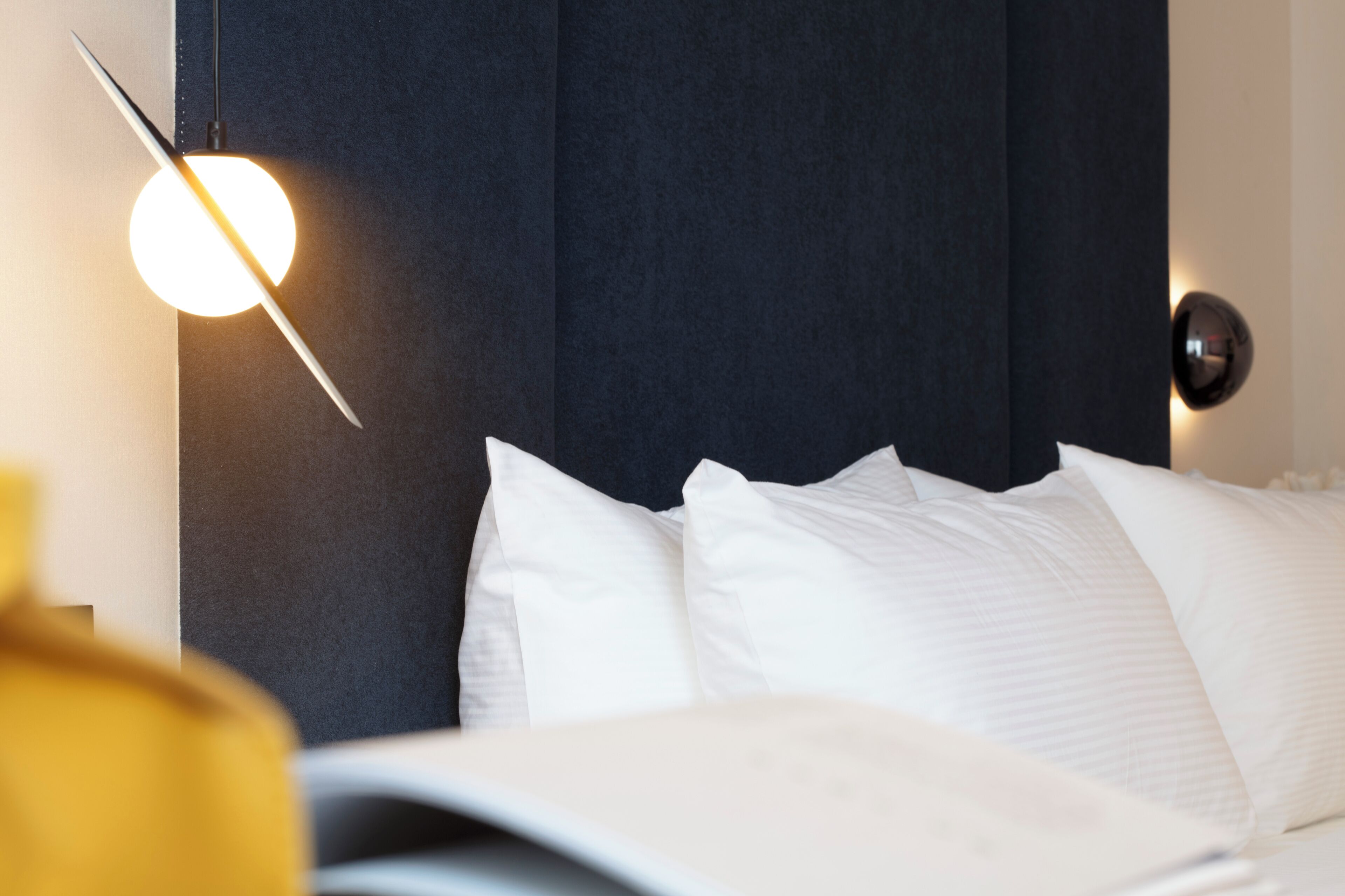 Una cama bien tendida con ropa blanca, cabecera oscura y una lámpara moderna fijada a la pared.