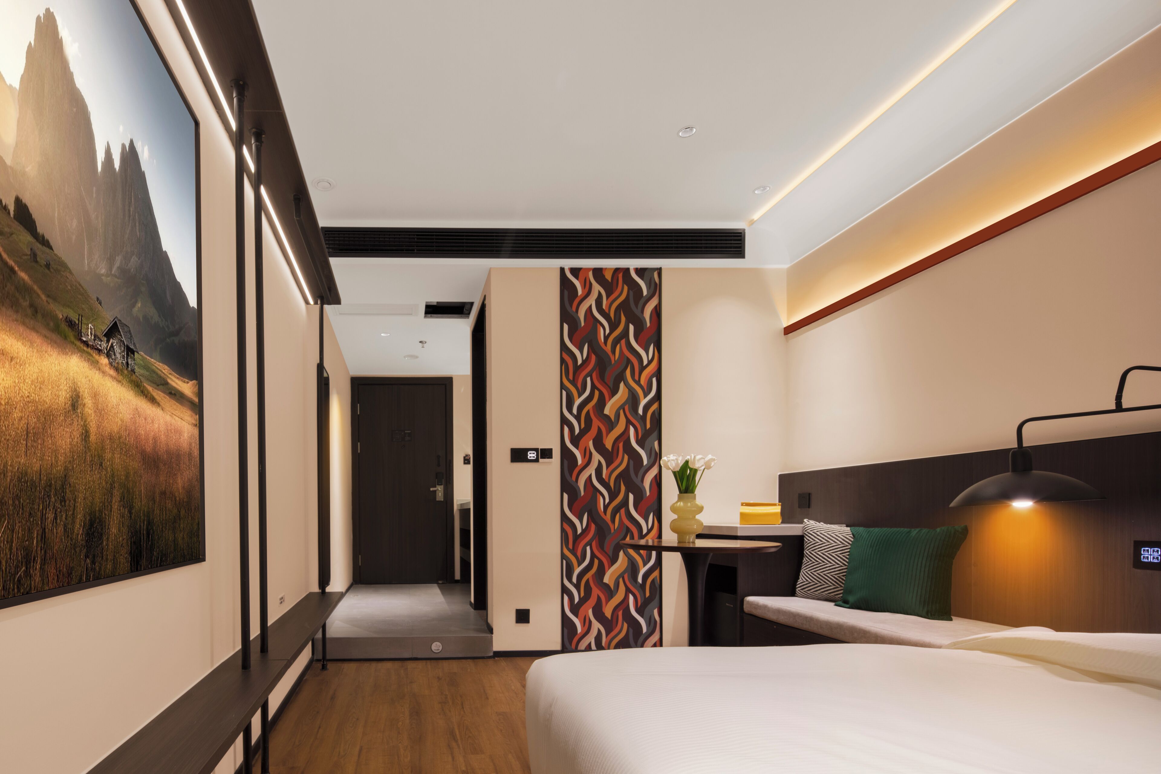 A modern hotel room with a large bed, a desk with a lamp, a view of mountains, and a patterned wall.