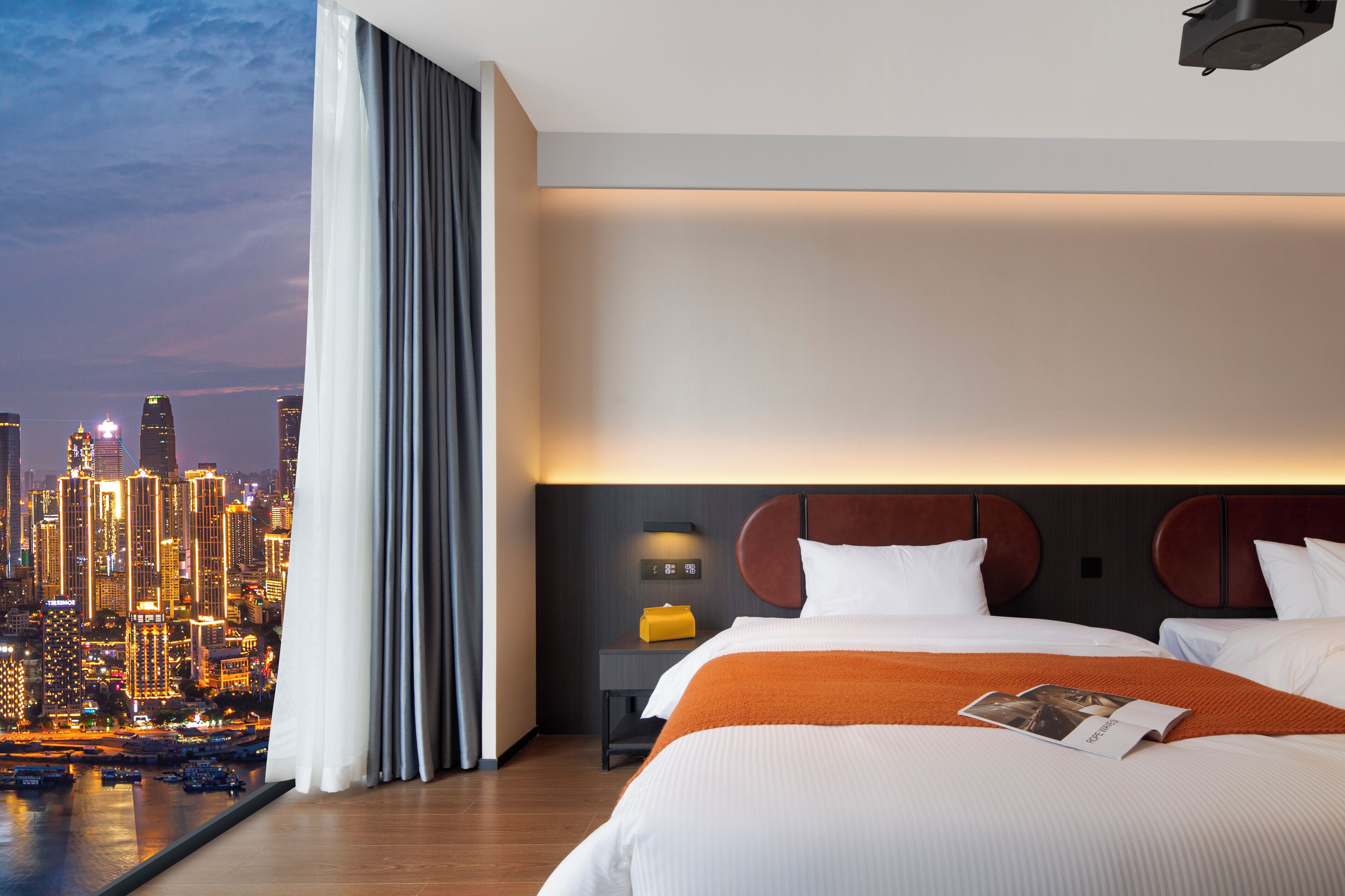 A hotel room with a city view, a large bed, a nightstand, and a modern headboard.