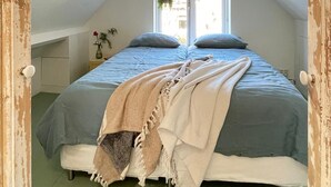 Una camera da letto con soffitto spiovente, una grande finestra, un letto con copriletto blu e una panca ai piedi del letto.