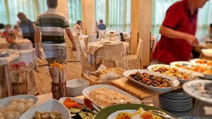 Uma mesa de buffet com diversos pratos, incluindo frutos do mar, legumes e saladas, em uma área de refeições bem iluminada, com mesas e cadeiras.