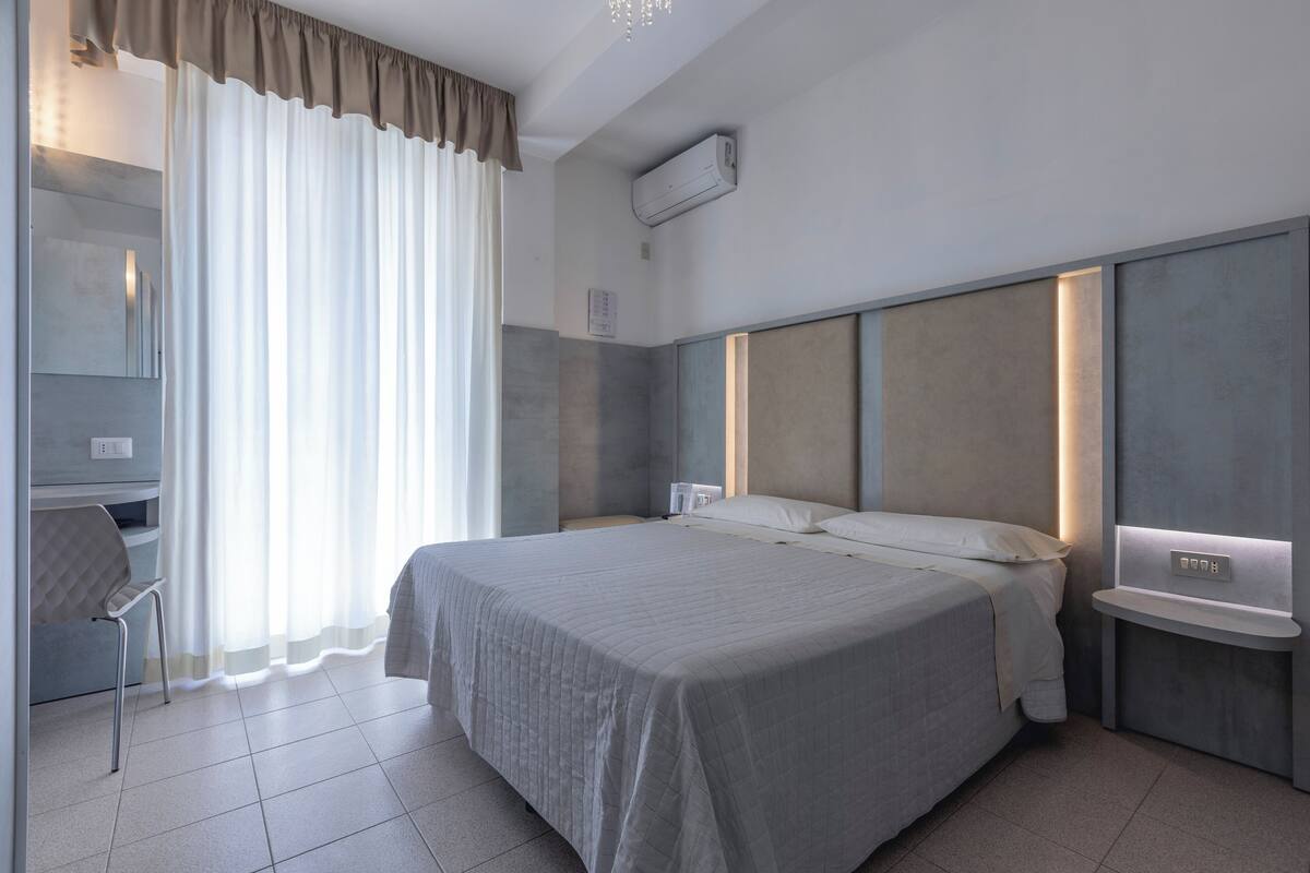 Moderni hotellihuone, jossa on suuri sänky, työpöytä ja tuoli. Huoneessa on ikkuna, jossa on verhot, ja seinällä on ilmastointilaite.