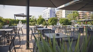 Espaço de pátio ao ar livre com mesas e cadeiras, cercado por guarda-sóis, com vista para um horizonte urbano com edifícios e áreas verdes.
