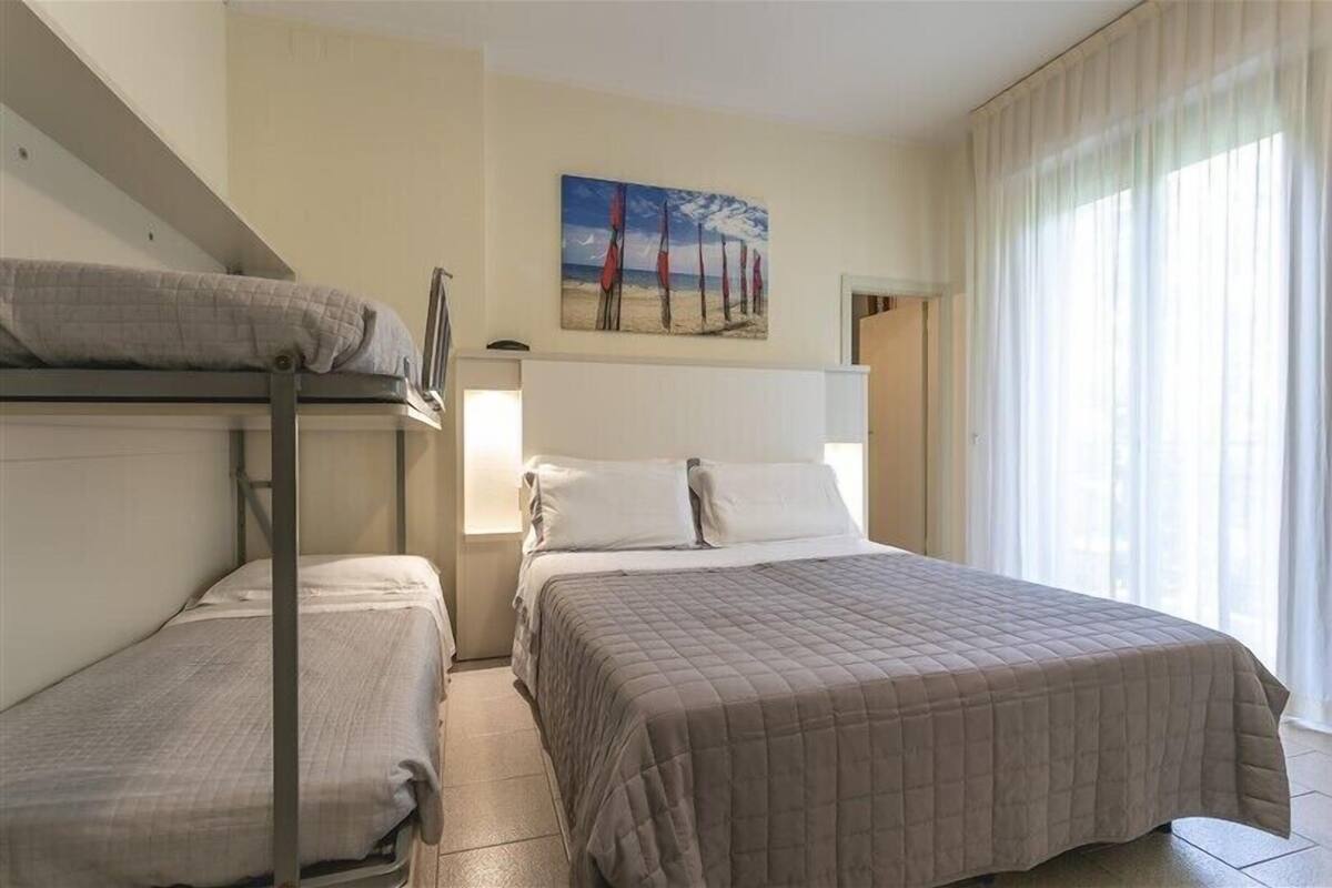 Hotellihuone, jossa on kerrossängyt, maalaus rantavarjoista ja suuri ikkuna, jossa on ohuet verhot.