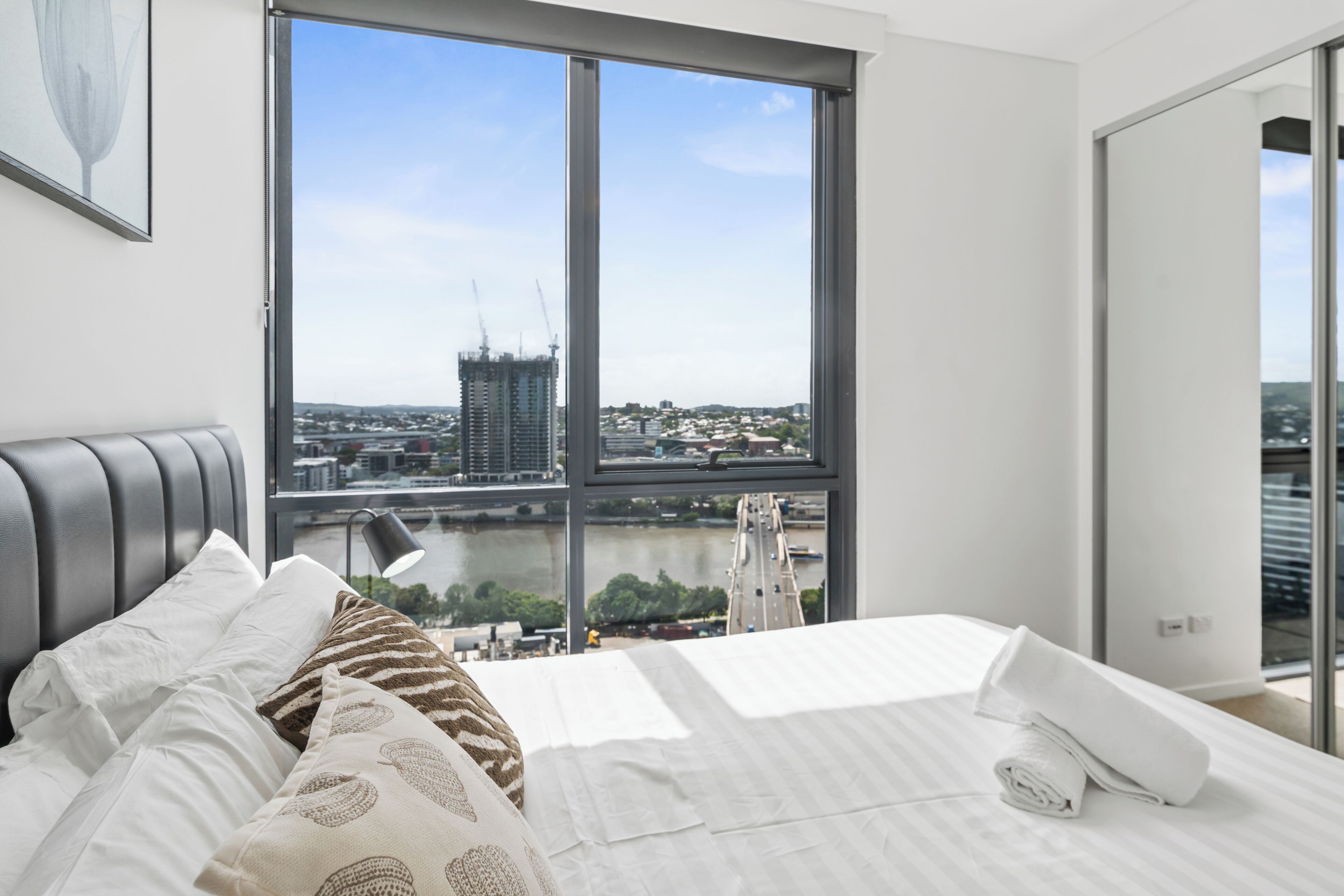 A modern bedroom with a large window, a bed with white linens, and a view of a cityscape.