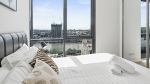 A modern bedroom with a large window, a bed with white linens, and a view of a cityscape. - Panorama river view with Free Parking (South Brisbane)