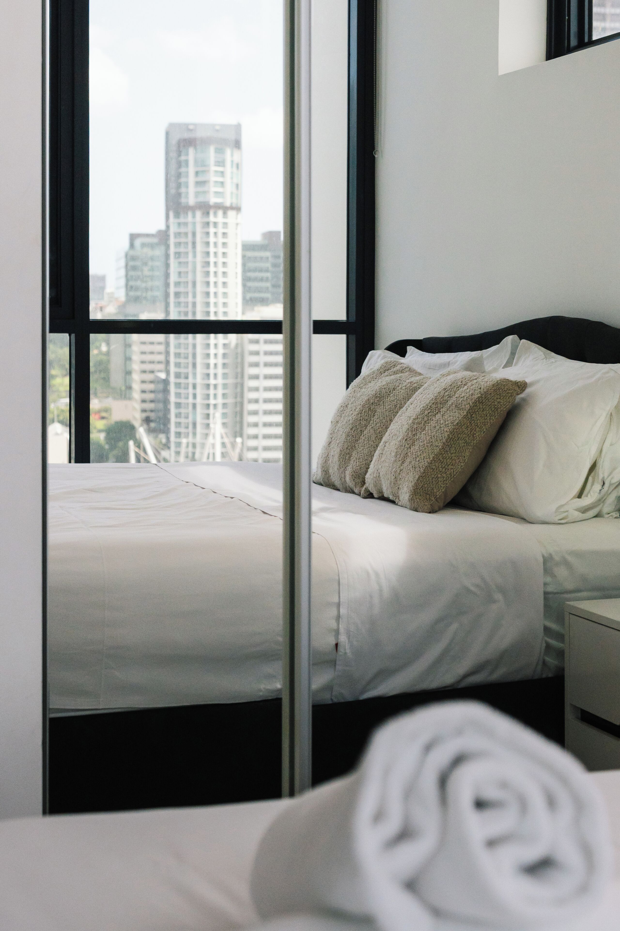 Ein Hotelzimmer mit einem Bett, Kissen, einem ordentlich bezogenen Bett, einem Fenster mit Blick auf hohe Gebäude und einem Nachttisch.