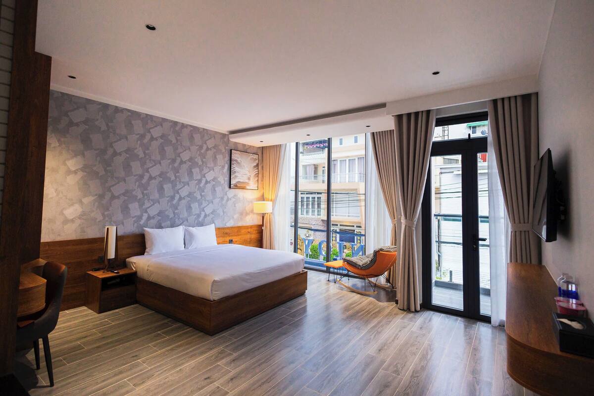 Habitación de hotel moderna con una cama grande, un escritorio, una silla, un televisor y un balcón con vistas a la ciudad.