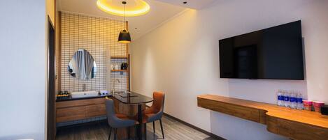 A modern hotel room with a wooden vanity, a round mirror, a dining table with two chairs, and a flat-screen TV mounted on the wall.
