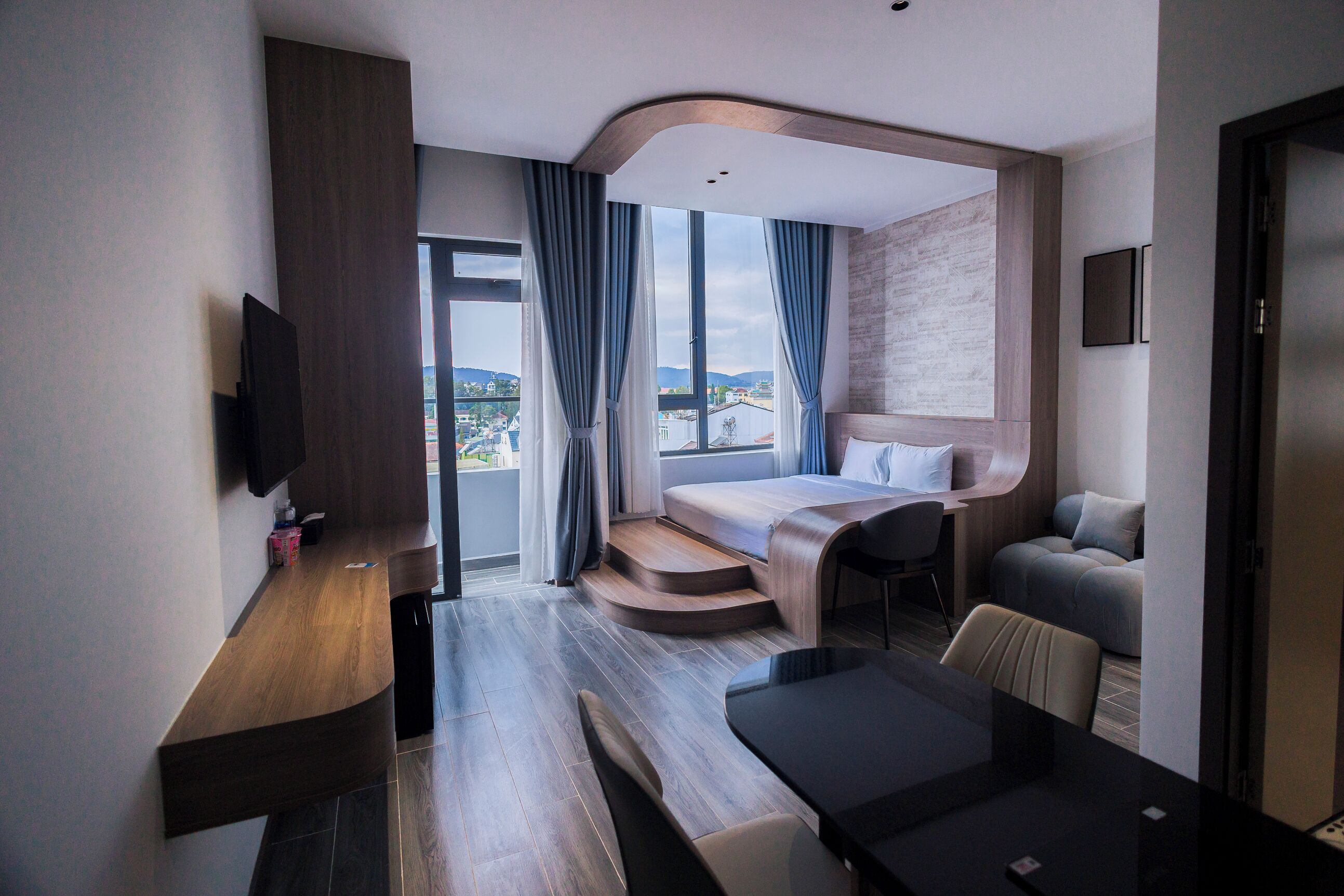 A modern hotel room with a curved desk, a bed with a view, a sofa, and a TV mounted on the wall.