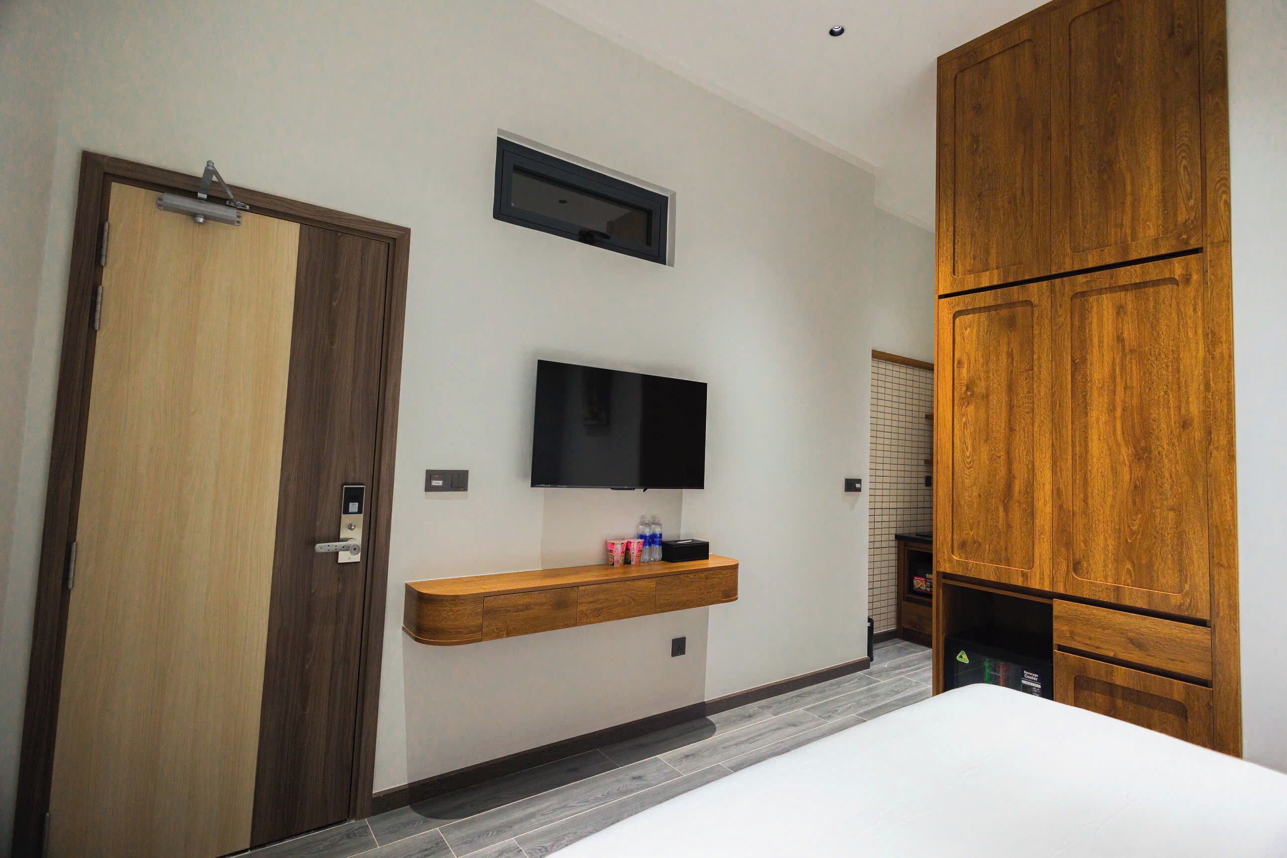 A modern hotel room with a wooden wardrobe, a mounted TV, and a built-in shelf with toiletries.