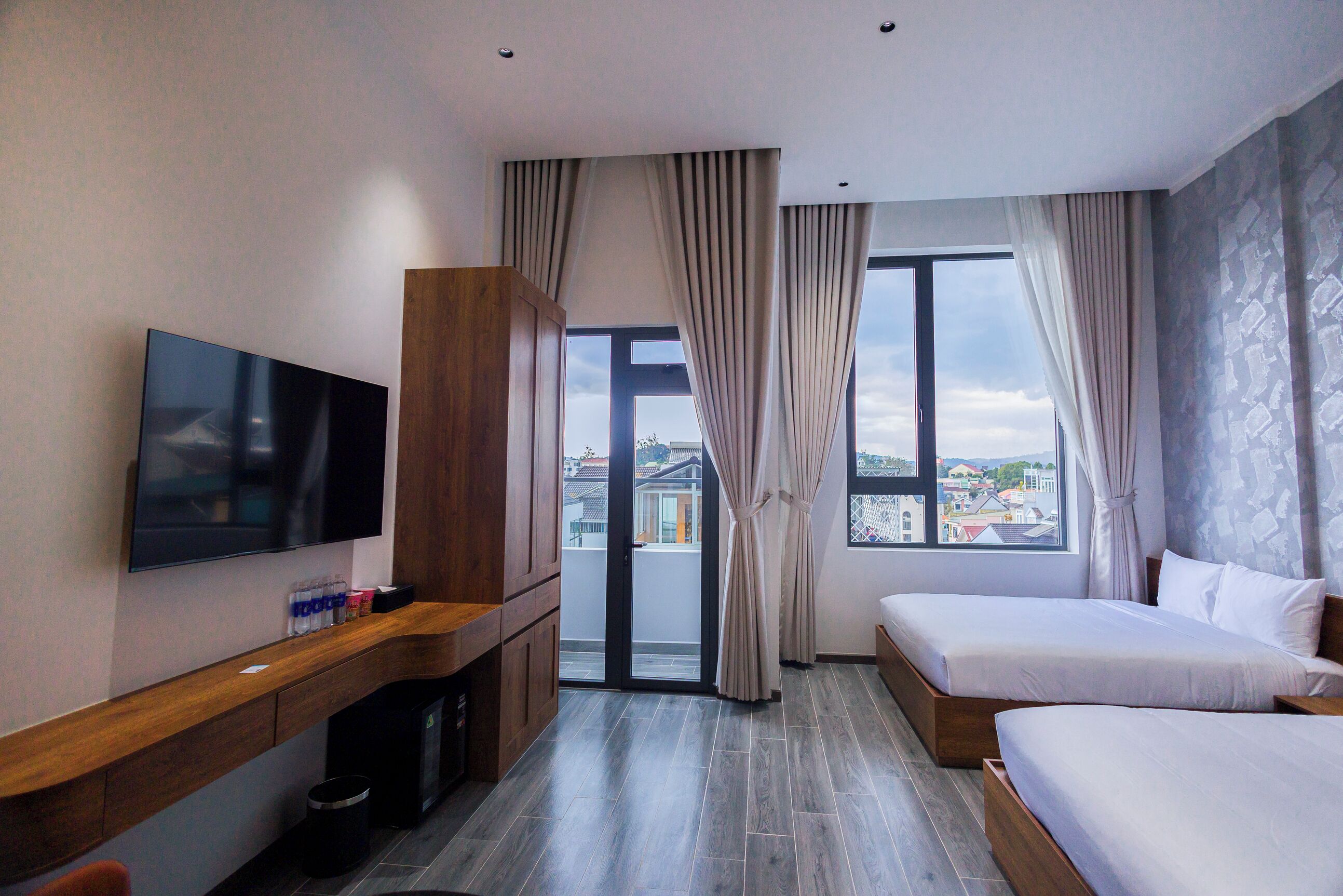 A hotel room with a TV, two beds, a wooden console, and a view of the city through large windows.