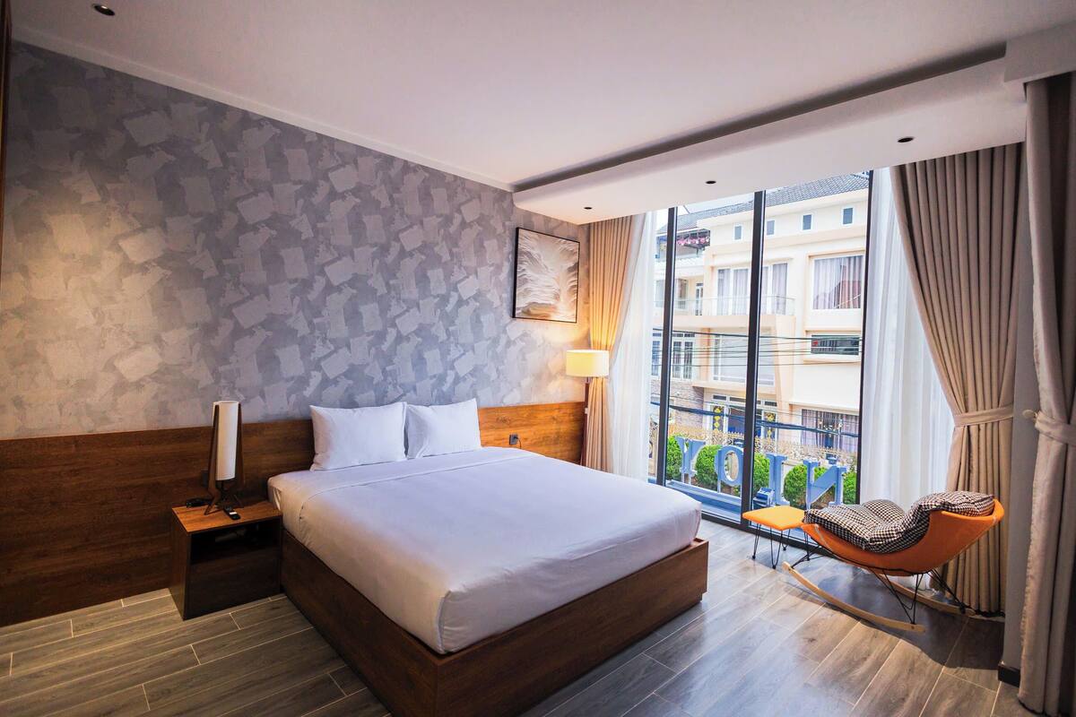 Una habitación de hotel moderna con una cama grande, una mesita de noche de madera, una mesita con lámpara, un sillón mecedora y vistas a la ciudad a través de amplios ventanales.