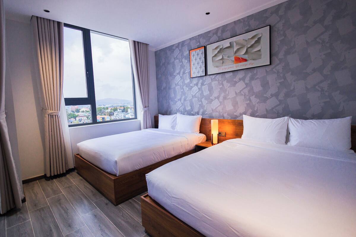 Habitación de hotel con dos camas, un amplio ventanal con cortinas, vista a la ciudad y dos cuadros en la pared.