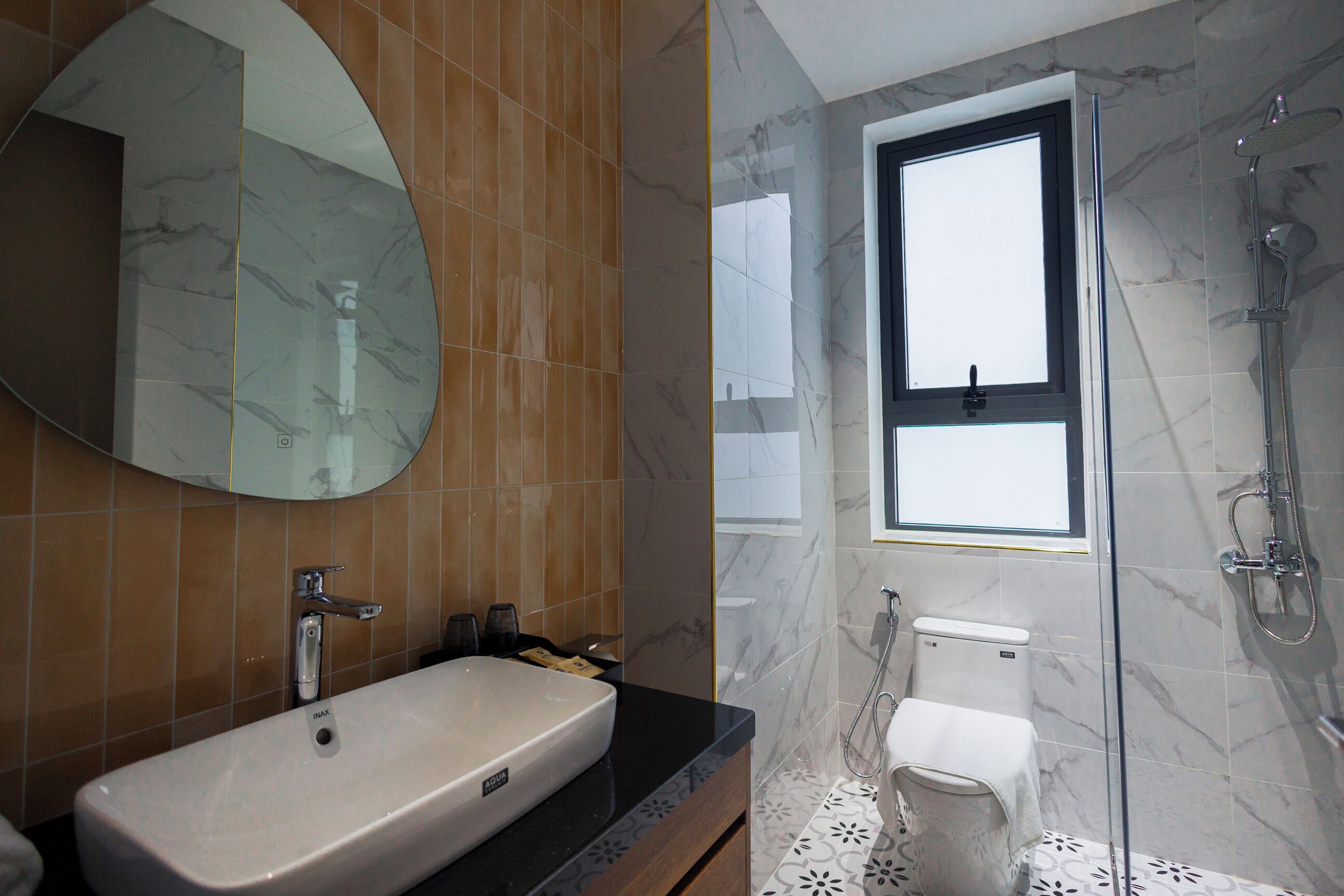 Et badeværelse med et stort ovalt spejl, en hvid vask, en bruseniche med glasdør og et vindue over toilettet.