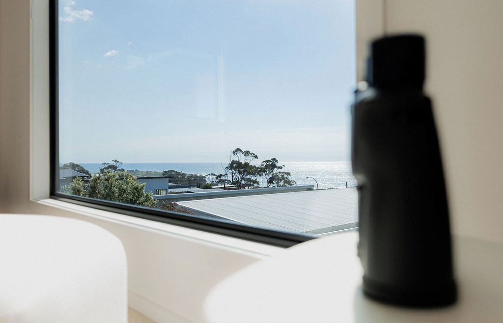 A room with a large window overlooking a coastal area, a black object on the windowsill, and a white chair in the foreground.