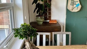 Een kamer met een bloempot op de vensterbank, een houten tafel met een boek, een groene deur en een tas aan de muur.