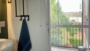 Een badkamer met een witte wastafel, een handdoekrek met een blauwe handdoek, een kamerplant op een plank, en uitzicht op een balkon met een hekwerk en een hanglamp.