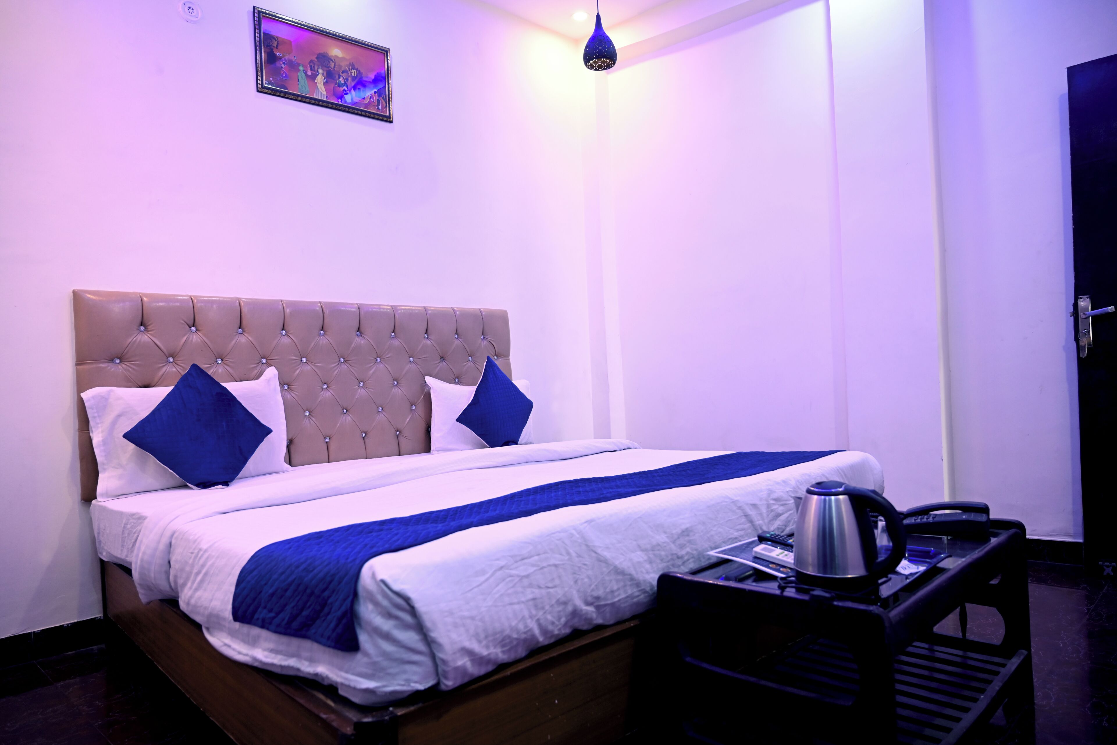 A neatly made bed with a tufted headboard, two pillows, and a blue and white bedspread. A small table with a kettle and a phone is beside the bed.