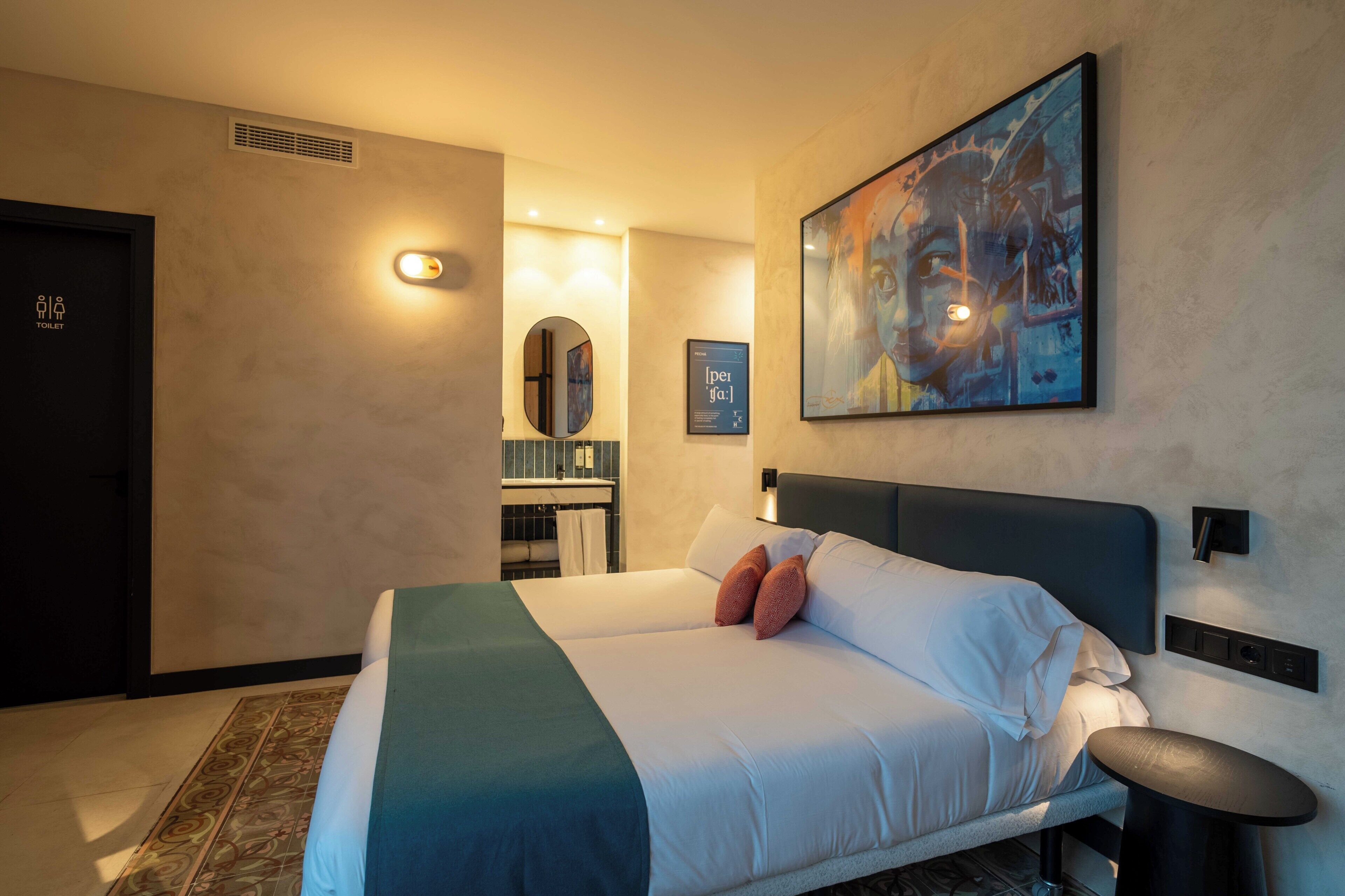 A hotel room with a bed, a bedside table, a wall-mounted light, a painting, a mirror, and a door with a toilet sign.