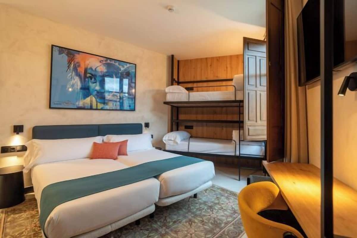 Une chambre d’hôtel avec un lit, un lit superposé, un bureau, une chaise, un tableau au mur et un tapis à motifs.