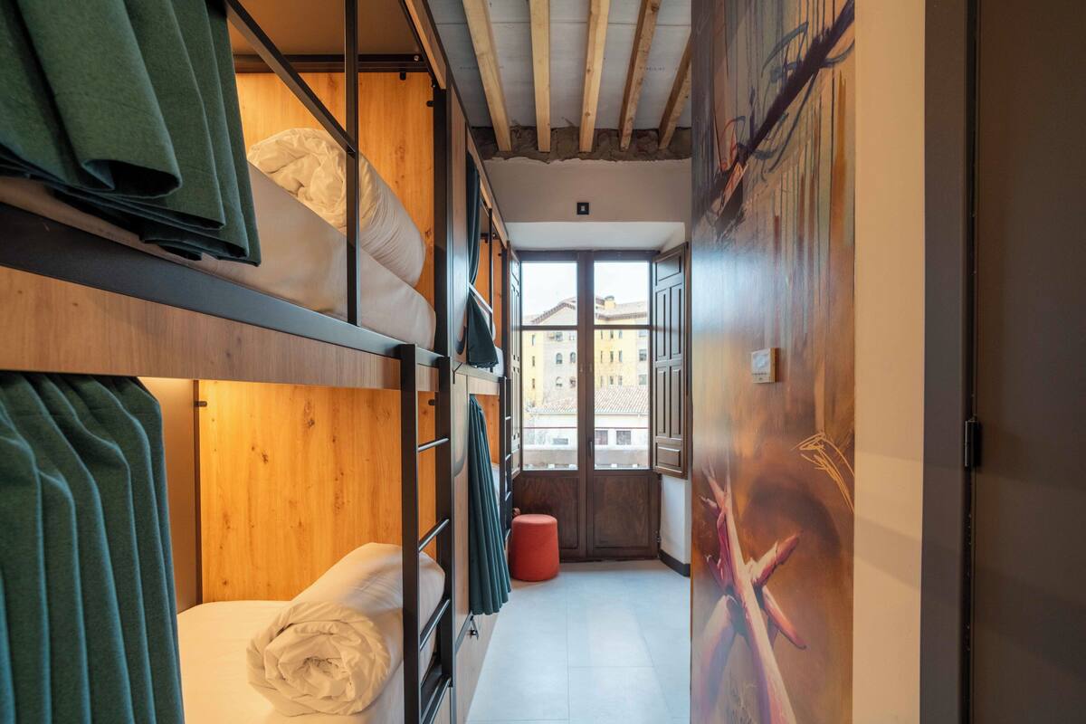 Un couloir étroit avec des lits superposés, une fenêtre donnant sur des immeubles et un tabouret rouge.