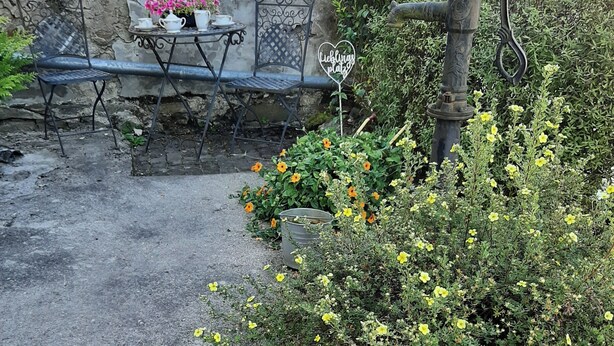 Een tuinpatio met een stenen muur, metalen stoelen, een tafel met theepotten, een hartvormig bordje en een waterkraan.