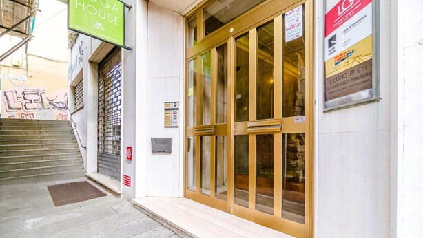 A building entrance with a glass door, a staircase leading to an upper floor, and a green sign above the door. - GuestHost - Porto Antico Apartment (Genova)