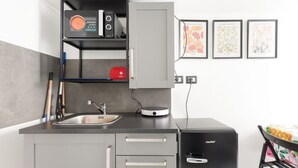 Eine kleine Küche mit Kaffeemaschine, Spüle und kleinem Kühlschrank. An den Wänden hängen Bilder in Rahmen und ein Stuhl mit einem blumigen Tischläufer.