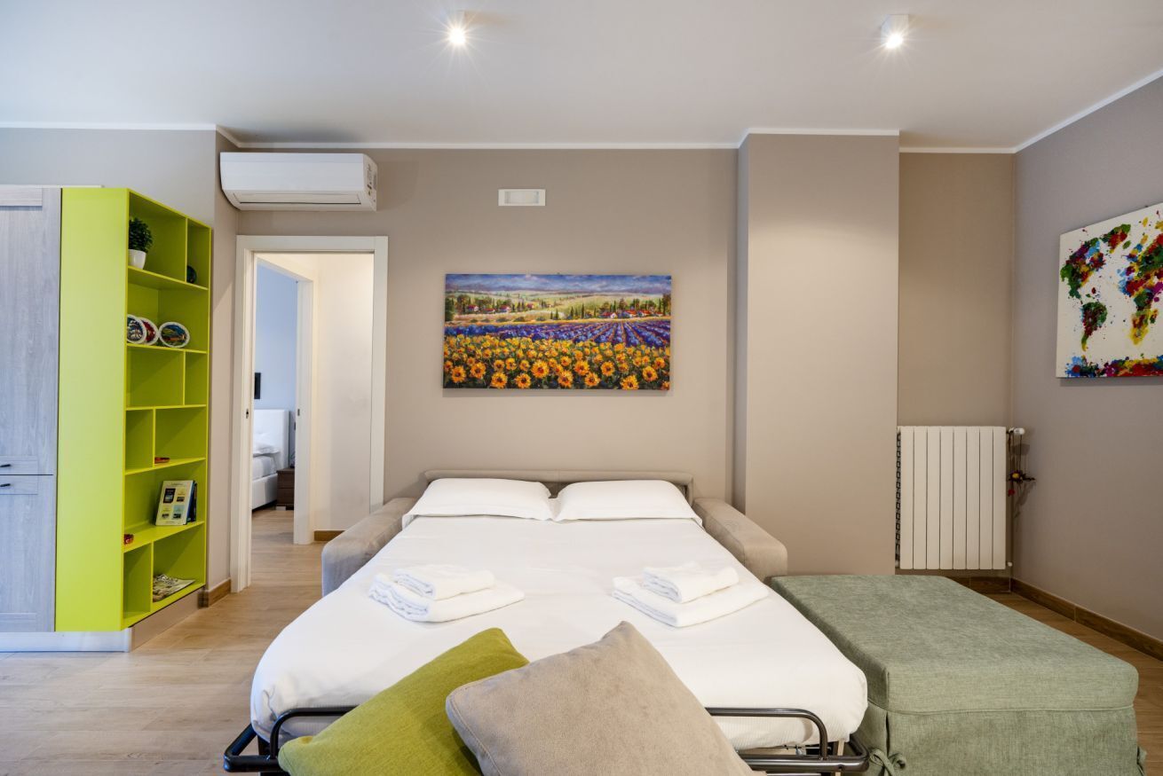 A hotel room with a bed, a green bookshelf, a painting of a sunflower field, and a radiator.