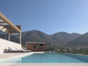 A modern outdoor pool area with lounge chairs and a wooden structure, surrounded by mountains. - Luxurious Villa Dias With Tennis Court by Estia (Mylopotamos)