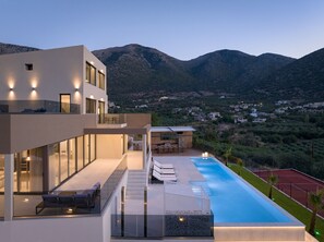 A modern multi-story building with a rooftop terrace, a swimming pool, and a view of a mountainous landscape. - Luxurious Villa Dias With Tennis Court by Estia (Mylopotamos)