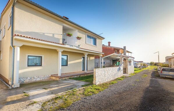 Un edificio residenziale a due piani con un balcone, un muro in pietra e un vialetto sterrato.
