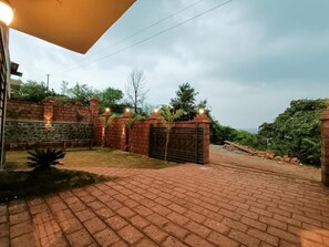 A paved driveway leading to a gated entrance with a stone wall and brick pillars, surrounded by greenery. - Omkar Villa (Moleshwar)