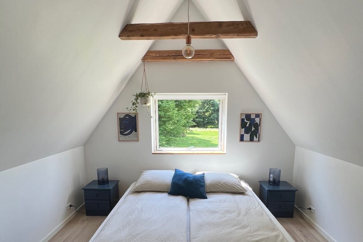 A bedroom with a sloped ceiling, a bed with a wooden headboard, a window with a view of greenery, and two bedside tables with lamps.