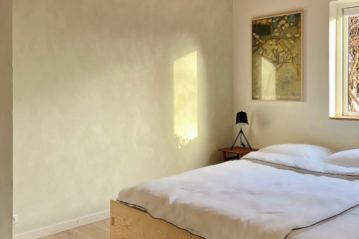 A bedroom with a wooden ceiling, a bed with white bedding, a small bedside table with a lamp, and a window with a view of trees.