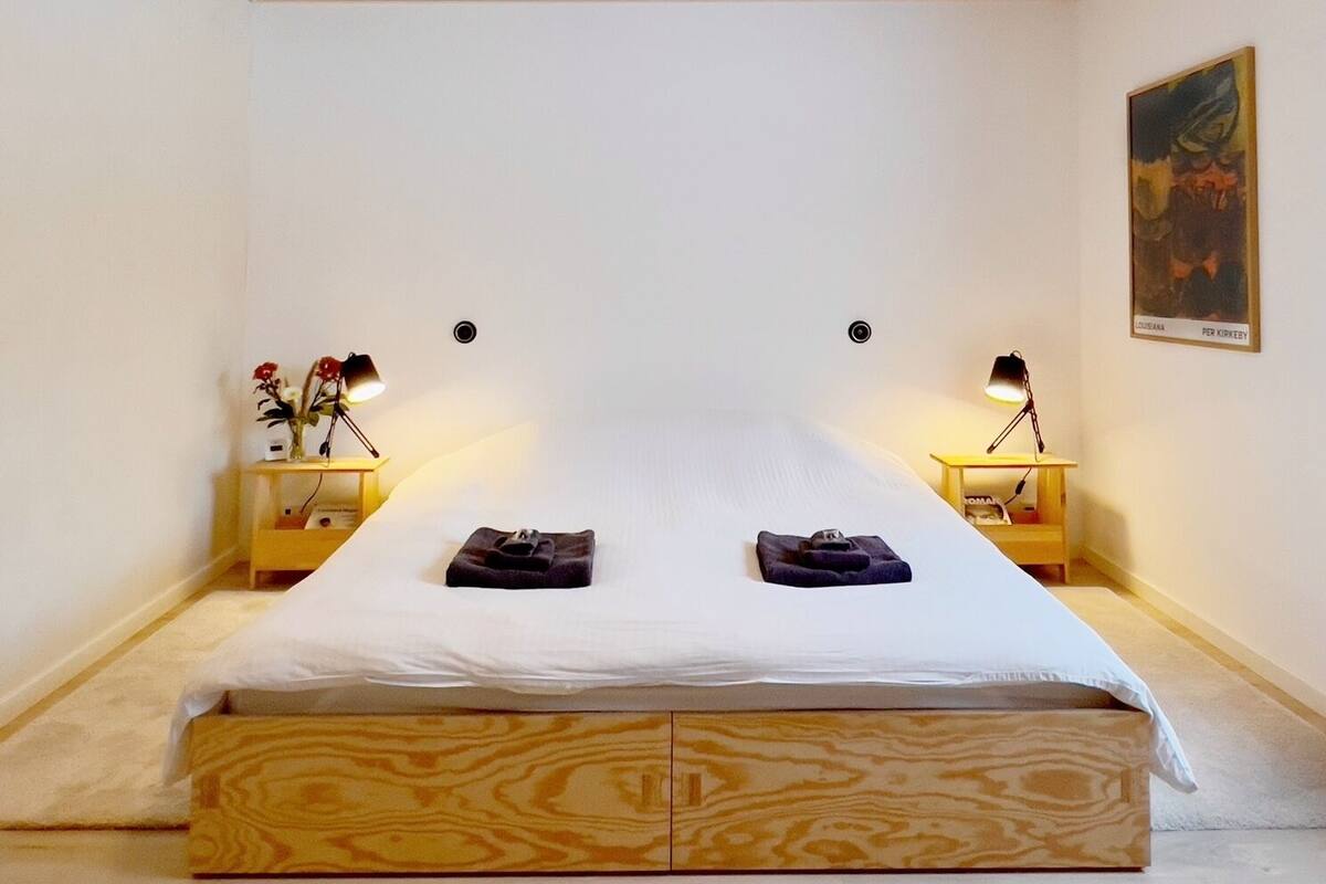 A small, cozy bedroom with a wooden ceiling, a single bed with a wooden headboard, two bedside tables with lamps, and a painting on the wall.