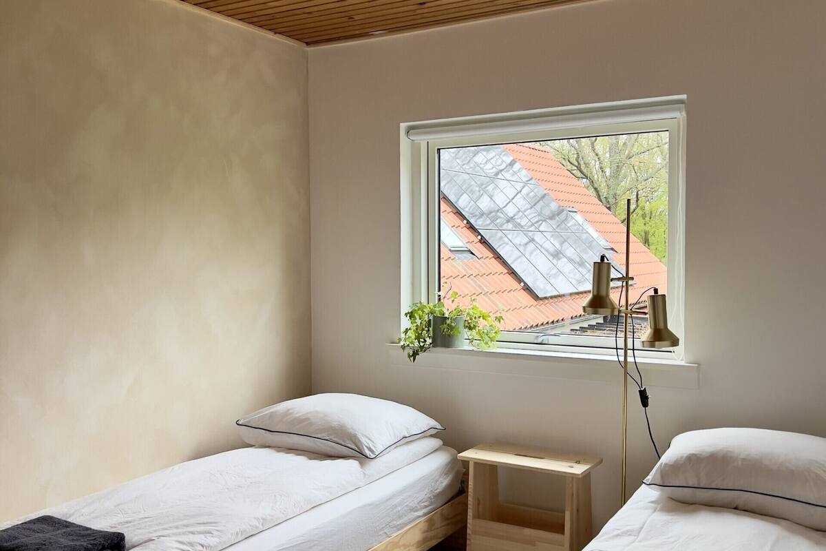 A room with two single beds, a small wooden nightstand, a window with a view of a building, and a ceiling with wooden slats.