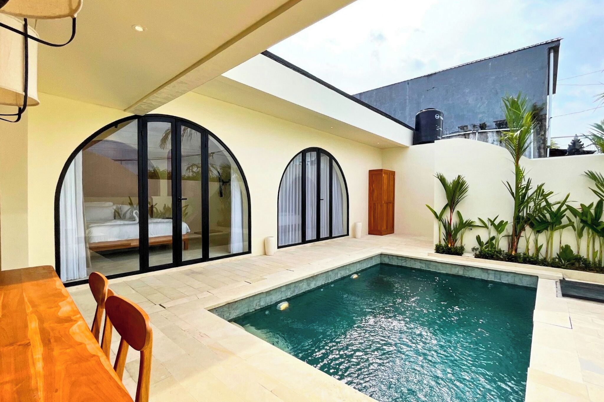 A modern outdoor pool area with a wooden table and chairs, a small garden with plants, and a large glass door leading to a bedroom.