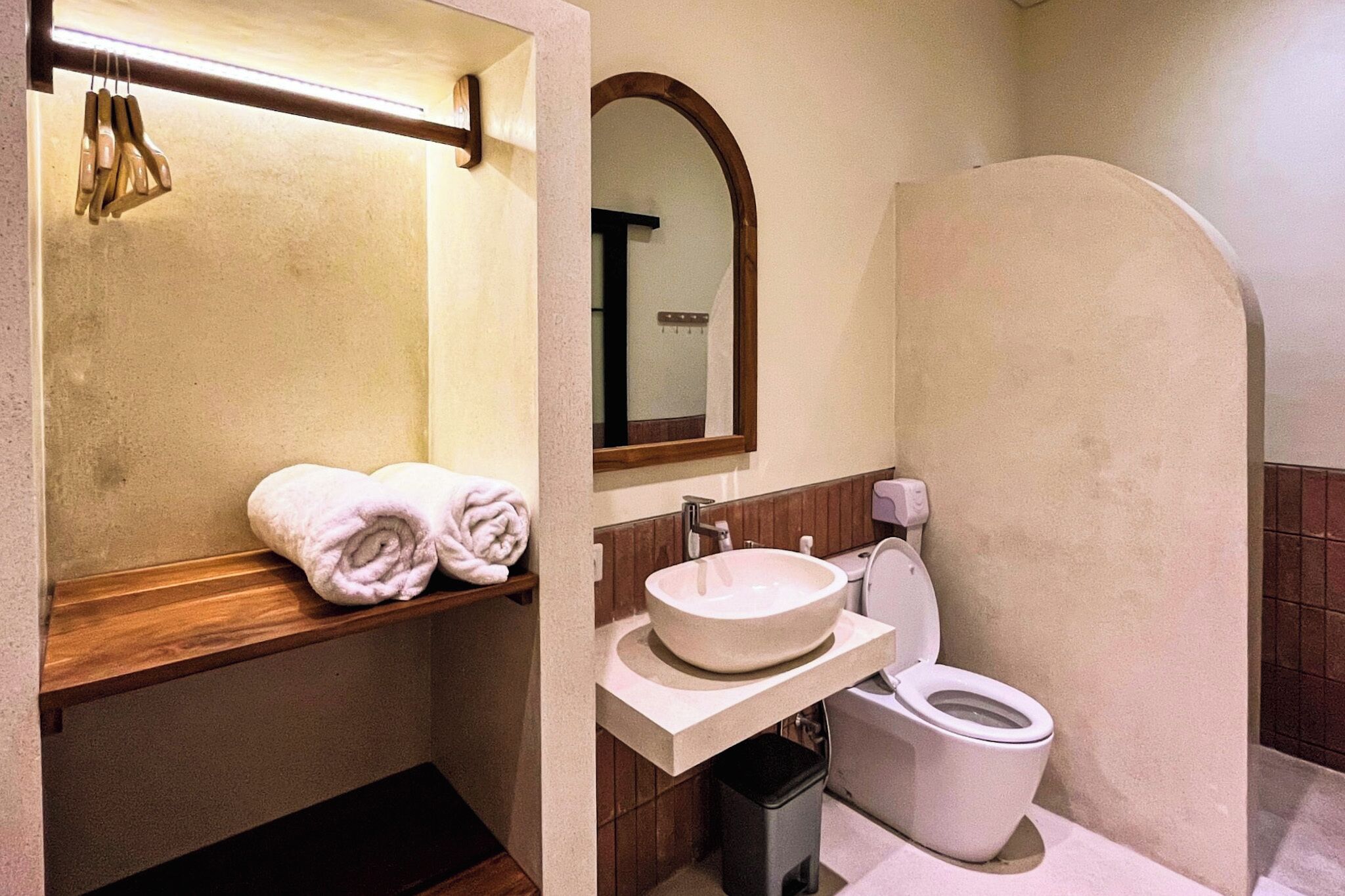 A bathroom with a wooden shelf holding towels, a sink with a mirror above it, and a toilet.