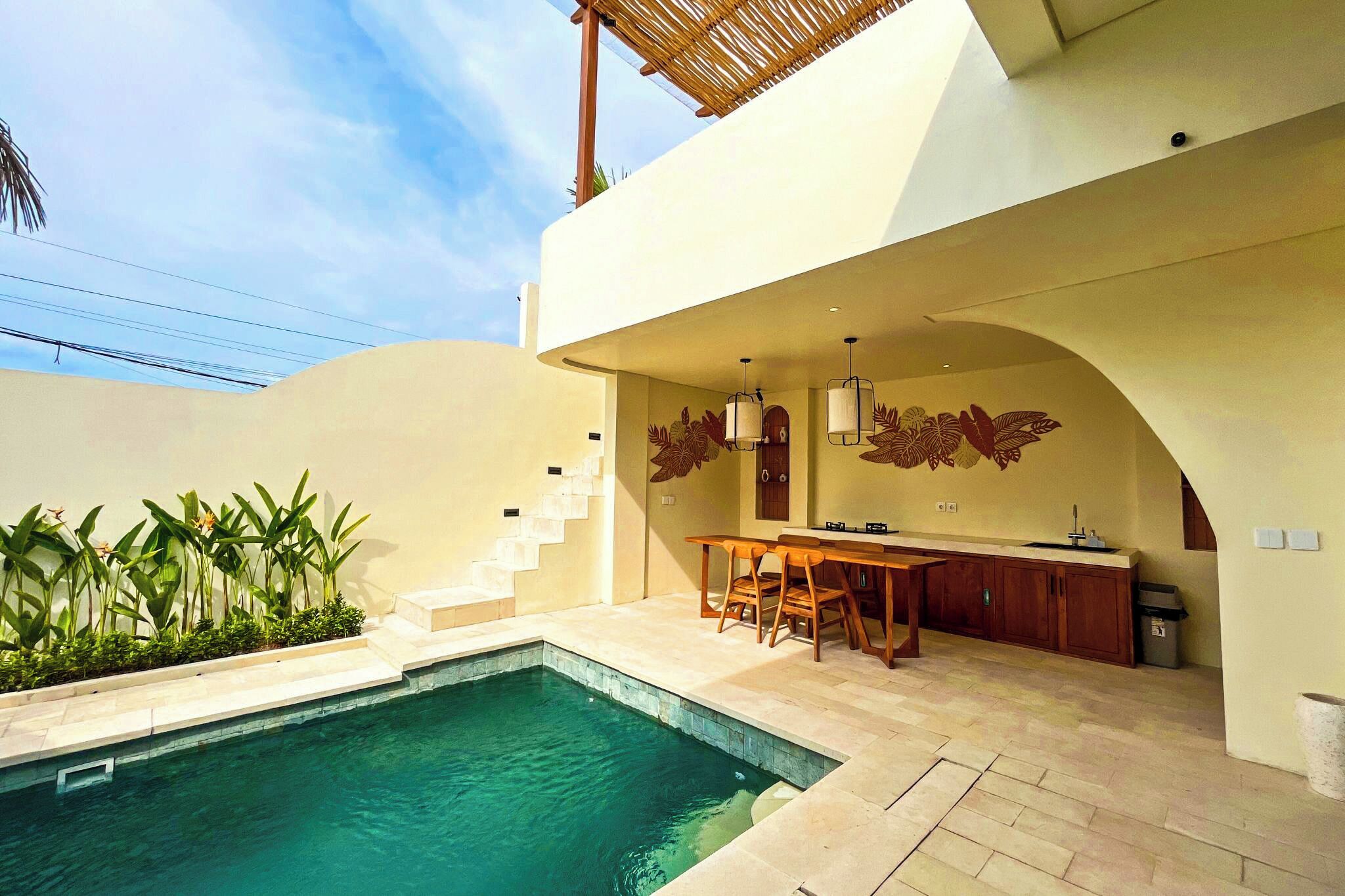 Un área de piscina moderna al aire libre con cocina y comedor, rodeada de plantas y una escalera.