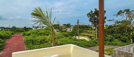 A balcony with a view of a red pathway leading to a lush green area with houses in the background.