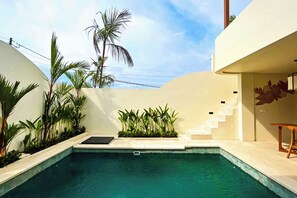 A swimming pool with a small staircase leading to a covered area, surrounded by lush greenery and a clear sky.