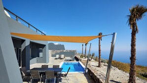 Area piscina moderna all'aperto con muro in pietra, tavolo e sedie, e vista sul mare.