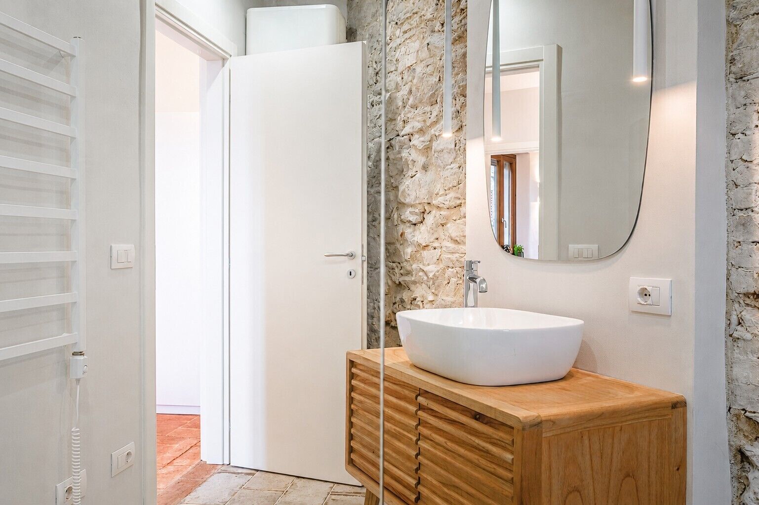 A bathroom with a white sink, wooden vanity, stone wall, and a door leading to another room.