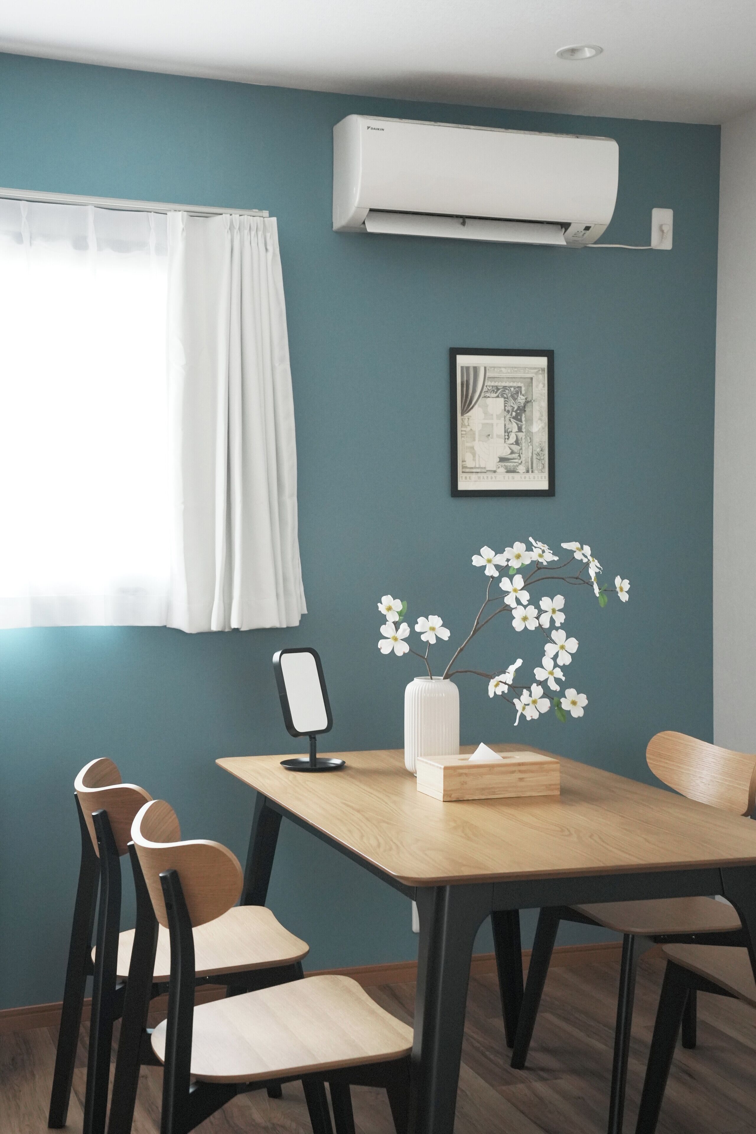 A dining area with a table, chairs, an air conditioner, a framed picture, and a vase with flowers.