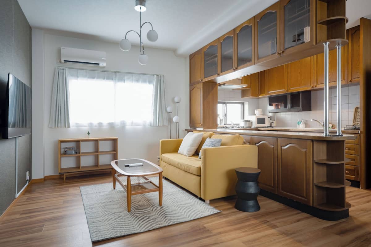 A modern living room with a sofa, a coffee table, and a TV. There is a kitchen with wooden cabinets and a dining area.