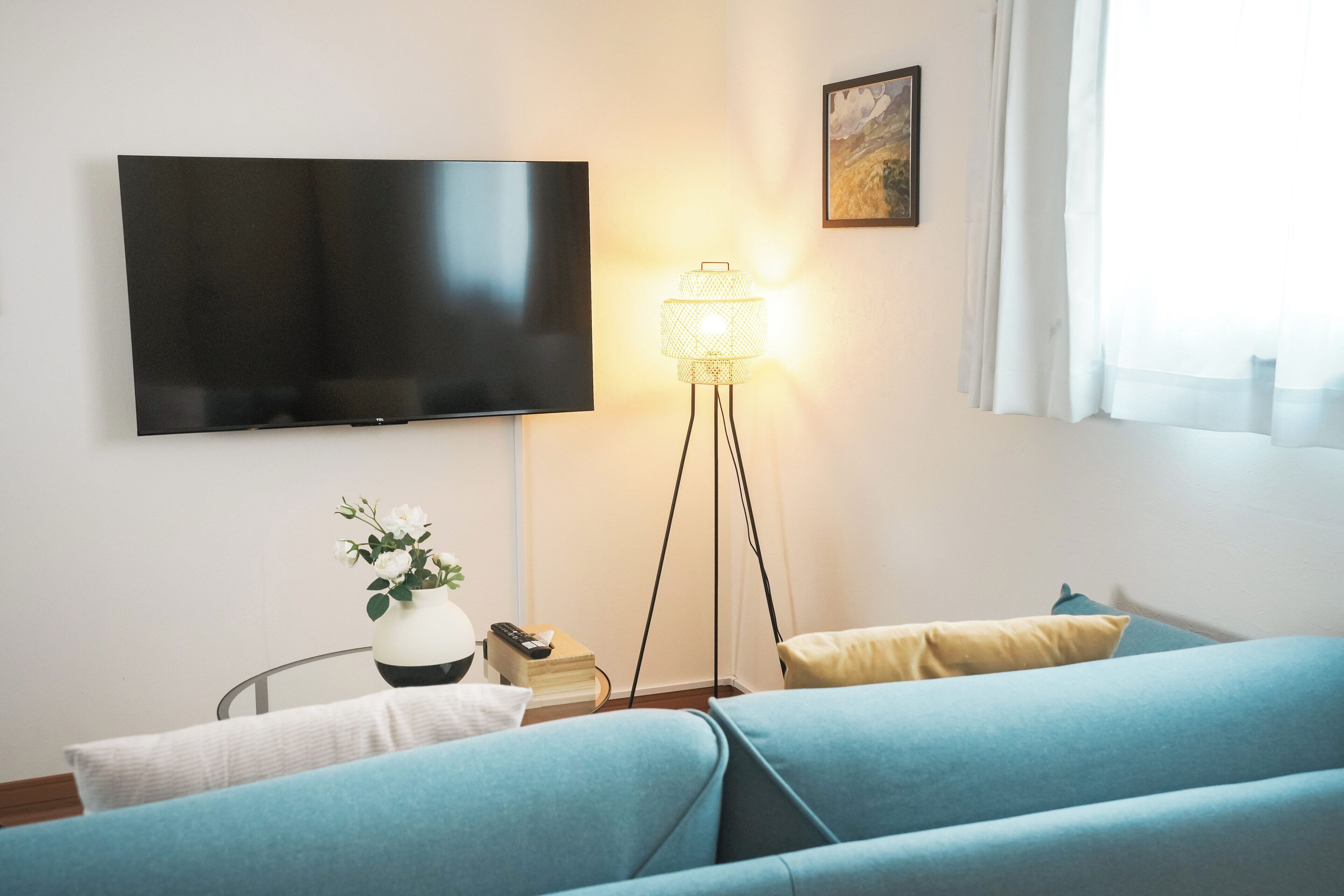 A hotel room with a flat-screen TV mounted on the wall, a floor lamp, a bed with blue bedding, and a bedside table with a vase of flowers.