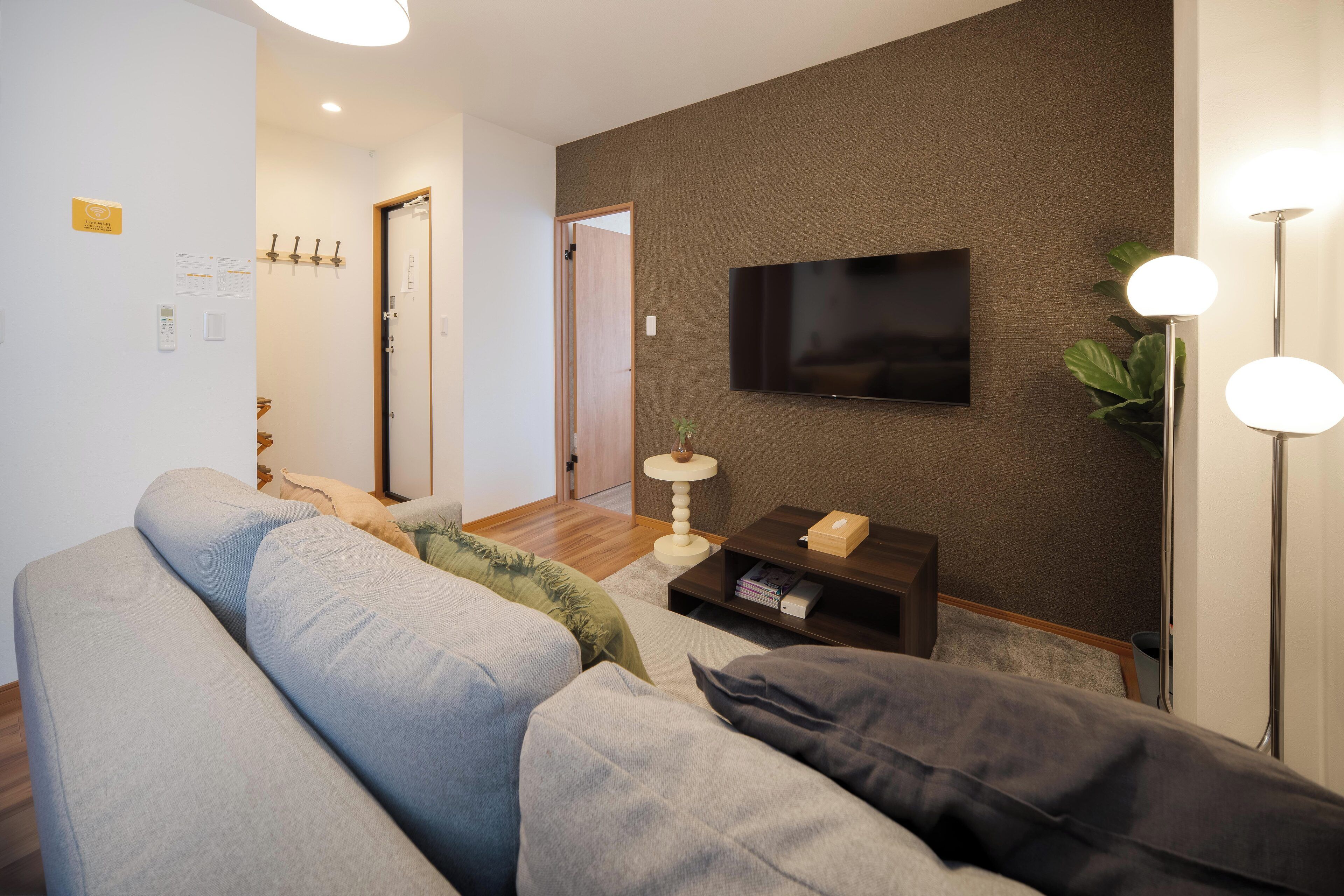 A modern living room with a grey sofa, a flat-screen TV mounted on the wall, a small side table with a lamp, and a wooden floor.